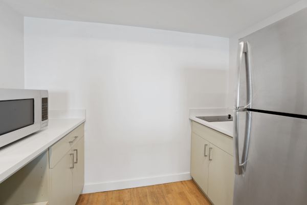 A small, modern kitchen with light wood flooring, white countertops, a microwave, cabinetry, a stainless steel refrigerator, and a small sink.