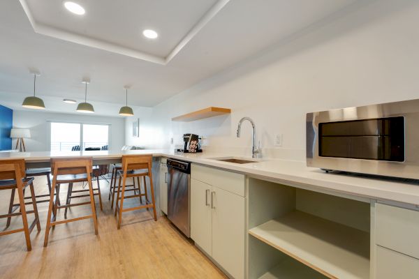 A modern kitchen with wooden floor, barstool seating, a microwave, an island, a sink, and pendant lights above an eating area.