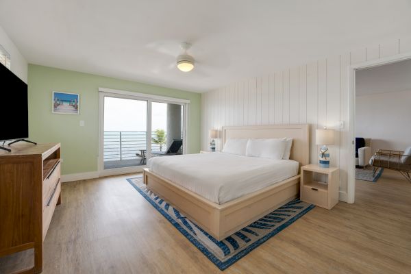 A spacious bedroom with a large bed, TV, light green accent wall, sliding glass doors to a balcony with a sea view, and an adjoining room entryway.