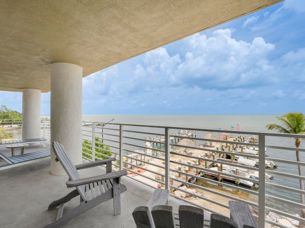 A balcony view with outdoor furniture overlooking the ocean, marina, and palm trees on a partly cloudy day, providing a serene atmosphere.