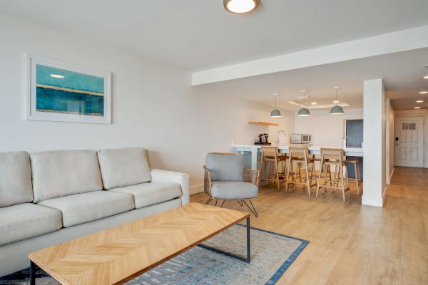 A modern living space with a sofa, armchair, coffee table, and an open kitchen area with barstools. The decor is minimalist and bright.
