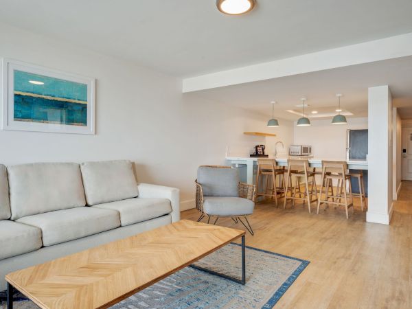 Bright modern living room with a sofa and coffee table, plus an open kitchen with bar stools and hanging pendant lights.