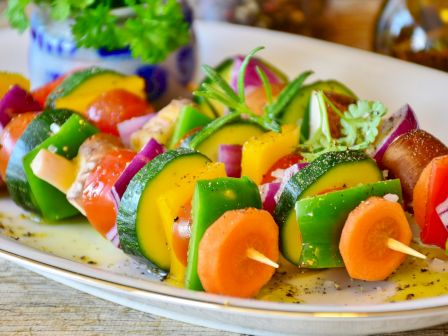 The image shows colorful vegetable skewers on a plate, including bell peppers, zucchini, onions, carrots, and other veggies, garnished with herbs.