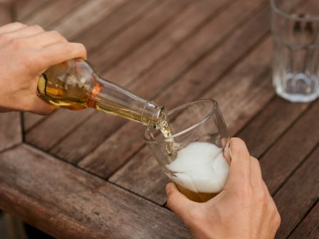 A person pours a drink from a bottle into a glass on a wooden table, with another empty glass in the background.