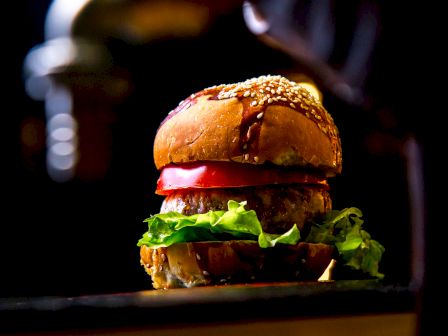 A gourmet burger with sesame seed bun, lettuce, tomato, and patty sits against a dark background, exuding a fresh and appetizing appearance.