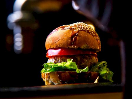A juicy sesame burger with lettuce and a red tomato slice, photographed up close against a dark background.