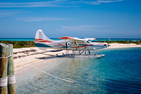 A seaplane is parked on a sandy beach near clear blue water, with a clear sky in the background.