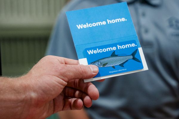 A person is holding a card with the text "Welcome home" and an image of a fish above another person's glimpse.