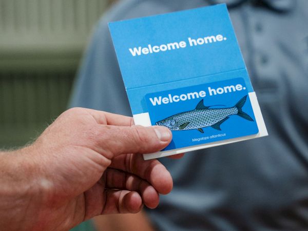 A person is holding a card with the text "Welcome home" and an image of a fish above another person's glimpse.