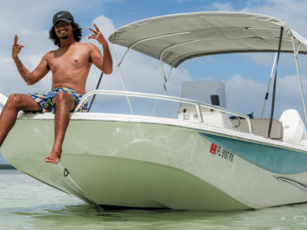 A shirtless individual in shorts poses on the bow of a white boat in shallow water, clear weather with an American flag on the boat’s stern.