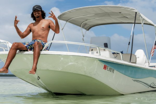 A shirtless individual in shorts poses on the bow of a white boat in shallow water, clear weather with an American flag on the boat’s stern.