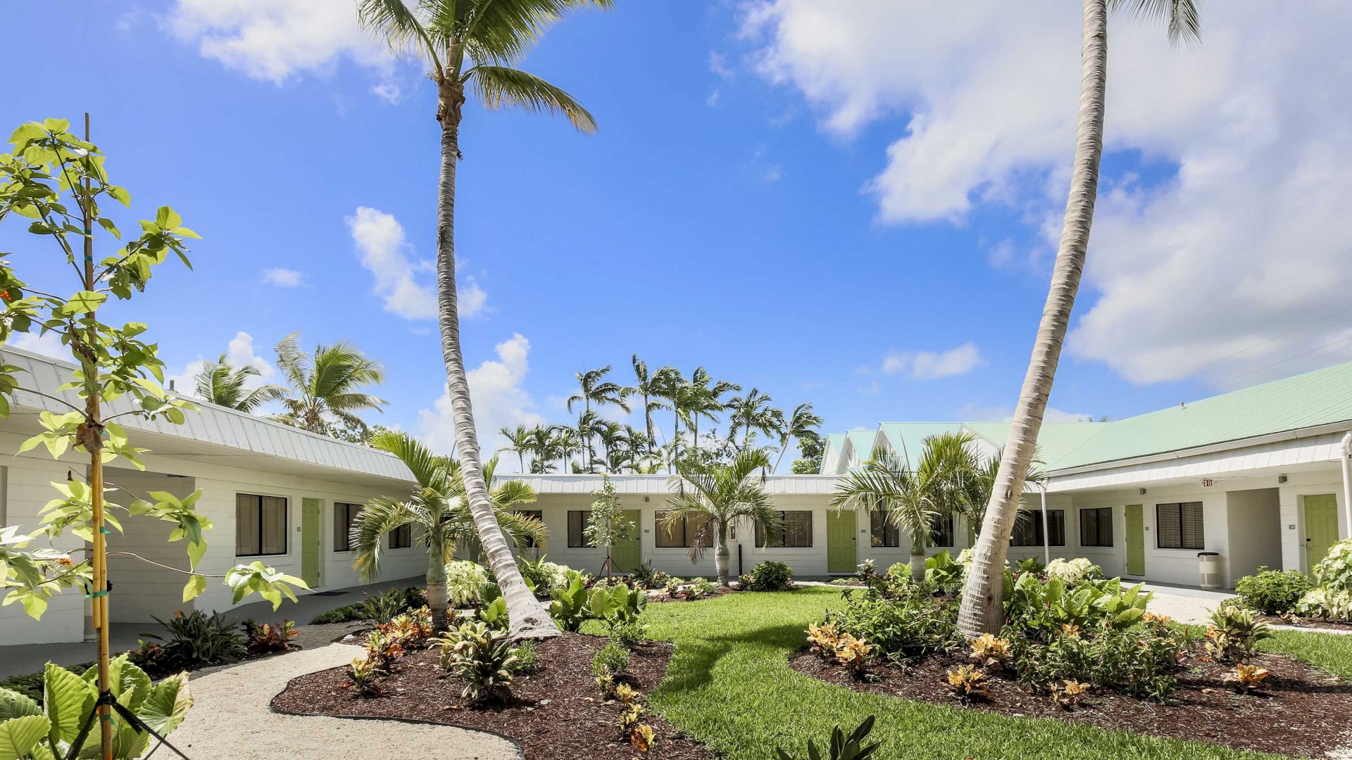 The image shows a courtyard with a garden, palm trees, and surrounding white buildings under a bright blue sky with scattered clouds.