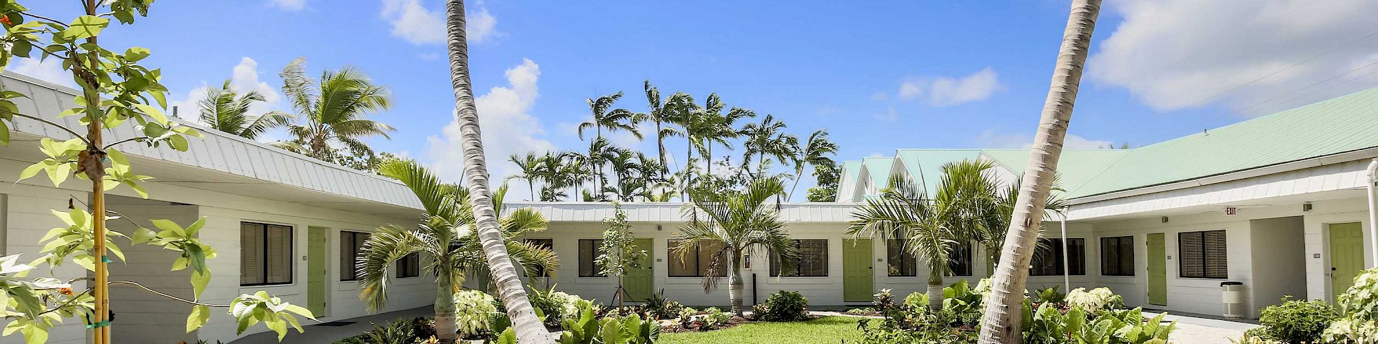 The image shows a courtyard with a garden, palm trees, and surrounding white buildings under a bright blue sky with scattered clouds.