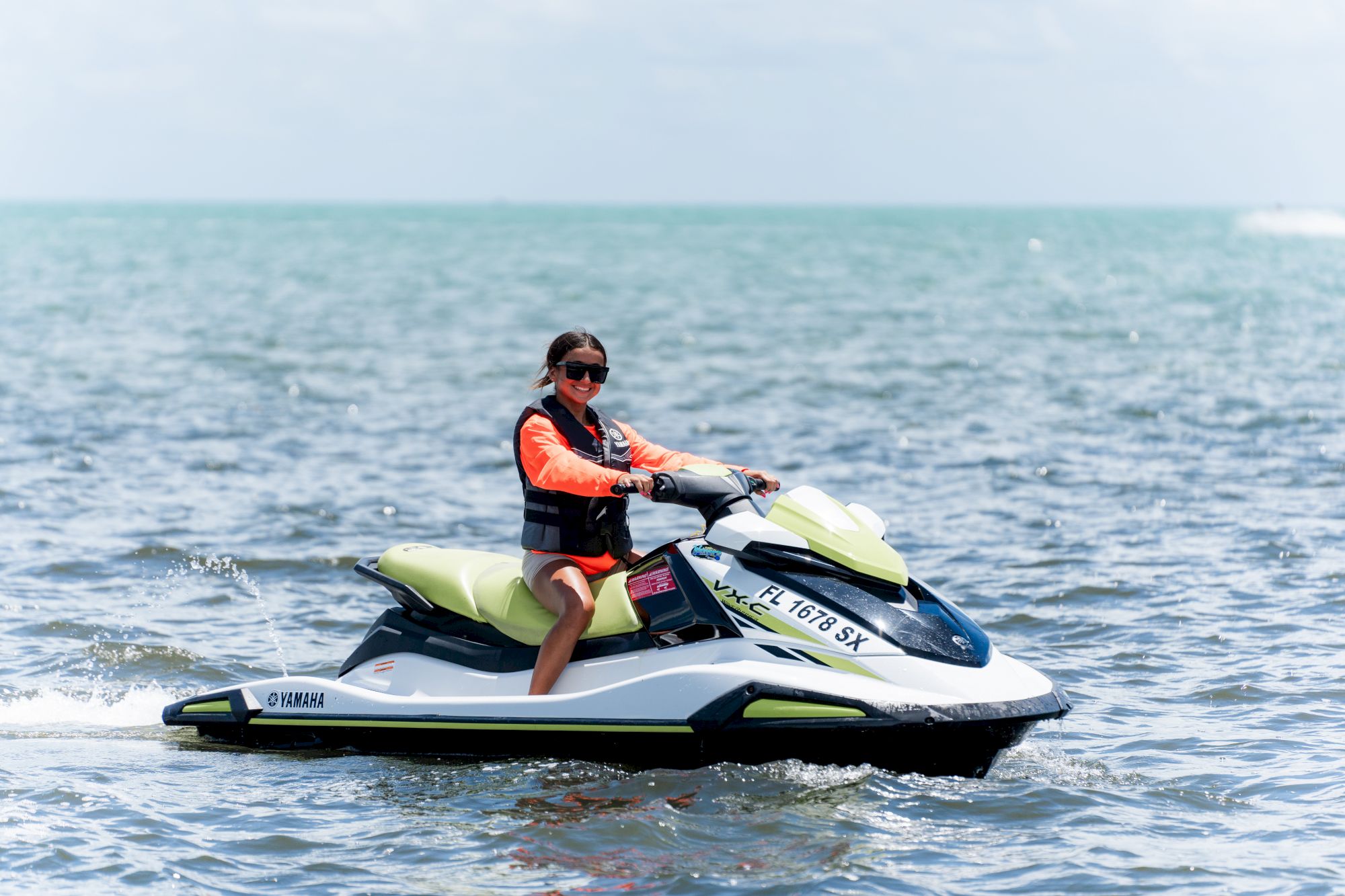 A person is riding a jet ski on a body of water. They are wearing a life jacket and sunglasses. The sky is cloudy in the background.