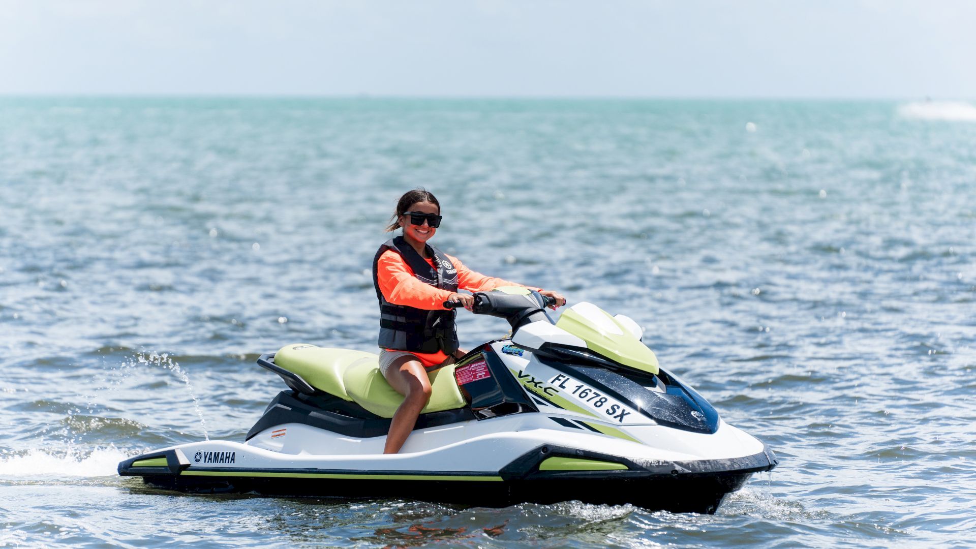 A woman in a life jacket smiles while riding a green-and-white jet ski across the ocean waves in sunny weather.