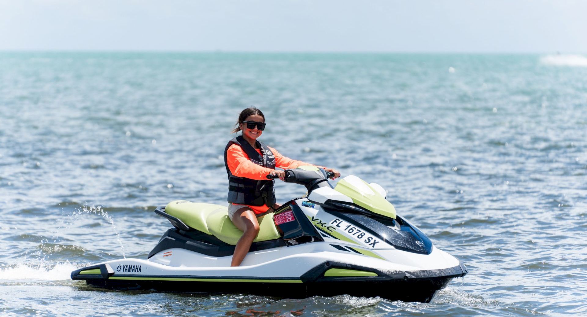 A woman in a life jacket smiles while riding a green-and-white jet ski across the ocean waves in sunny weather.