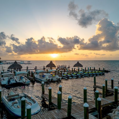 The image shows a picturesque sunset over a marina with boats docked at the wooden piers and huts in the water, under a sky with scattered clouds.