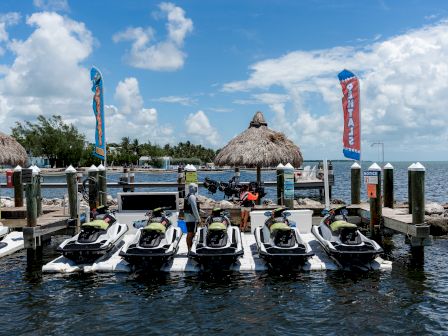 A dock with several jet skis lined up, two huts, and flag banners near a body of water with a clear blue sky.