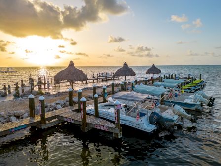 Boats are docked at a marina during sunset, with thatched-roof huts in the background and the ocean stretching to the horizon.