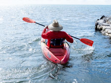 A person in a red kayak paddles away from a rocky shoreline into the open water under a clear blue sky, wearing a wide-brim hat.