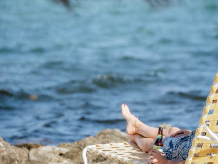 A person is relaxing on a lounge chair by the beach, legs extended, with the ocean and rocky shore in the background.