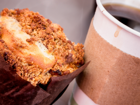 A close-up of a muffin with a crispy topping next to a disposable coffee cup wrapped in a cardboard sleeve on a table.