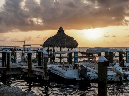 Boats are docked at a pier as the sun sets over the ocean, with a thatched-roof structure in view and clouds filling the orange sky in the background.