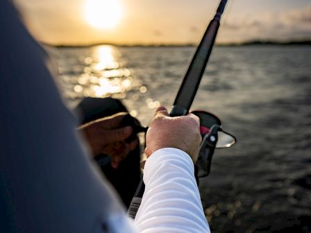 A person aboard a boat fishes at sunset, holding a rod with a calm water backdrop and warm sky, capturing a peaceful outdoor moment.