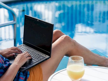 A person is using a laptop by a pool while sitting on a lounge chair, with a glass of juice on a small table nearby.