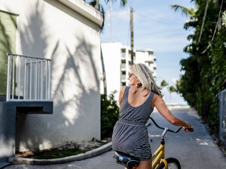 A person with blonde hair in a gray dress is riding a yellow bicycle on a sunny street lined with palm trees and buildings.