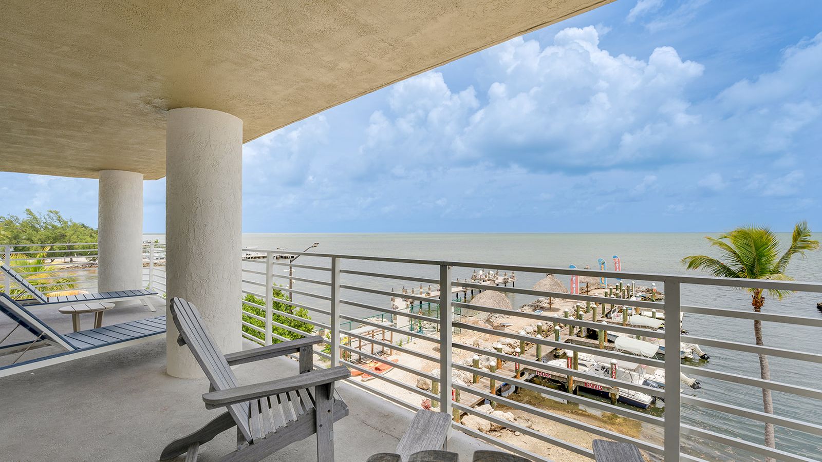 The image shows a balcony with chairs overlooking a marina and the ocean under a cloudy sky. There are boats and a pier visible below.