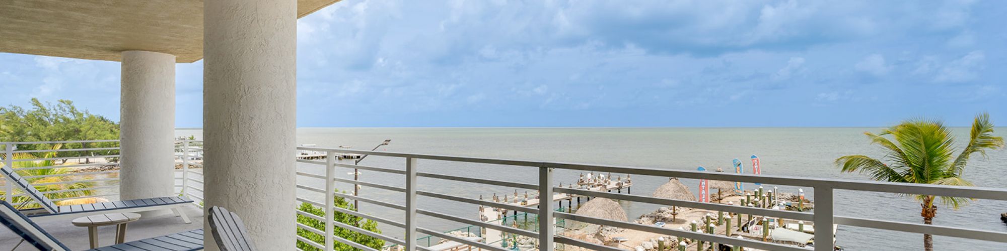 The image shows a balcony with chairs overlooking a marina and the ocean under a cloudy sky. There are boats and a pier visible below.