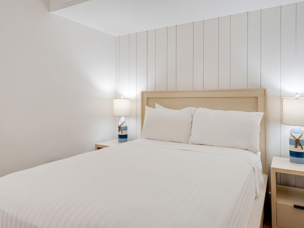 A bedroom with a neatly made bed, white linens, two bedside tables with matching lamps, and a white shiplap accent wall.