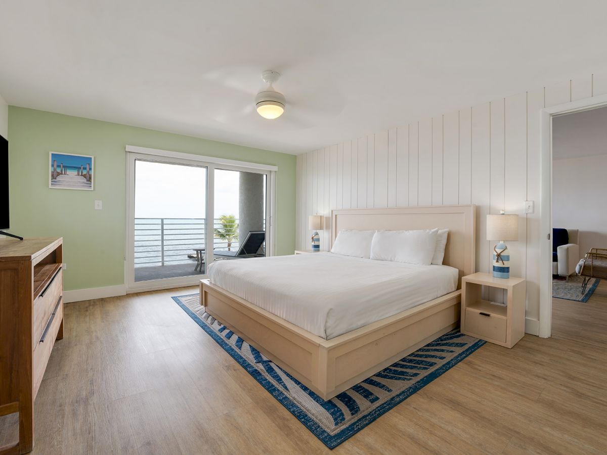 A modern bedroom with a large bed, TV, wooden floor, and a balcony with ocean views. There's a doorway leading to another room.