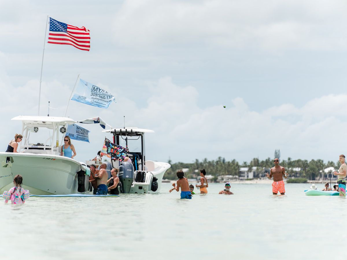 People swim near a white speedboat in a sunny bay as a large American flag waves overhead.