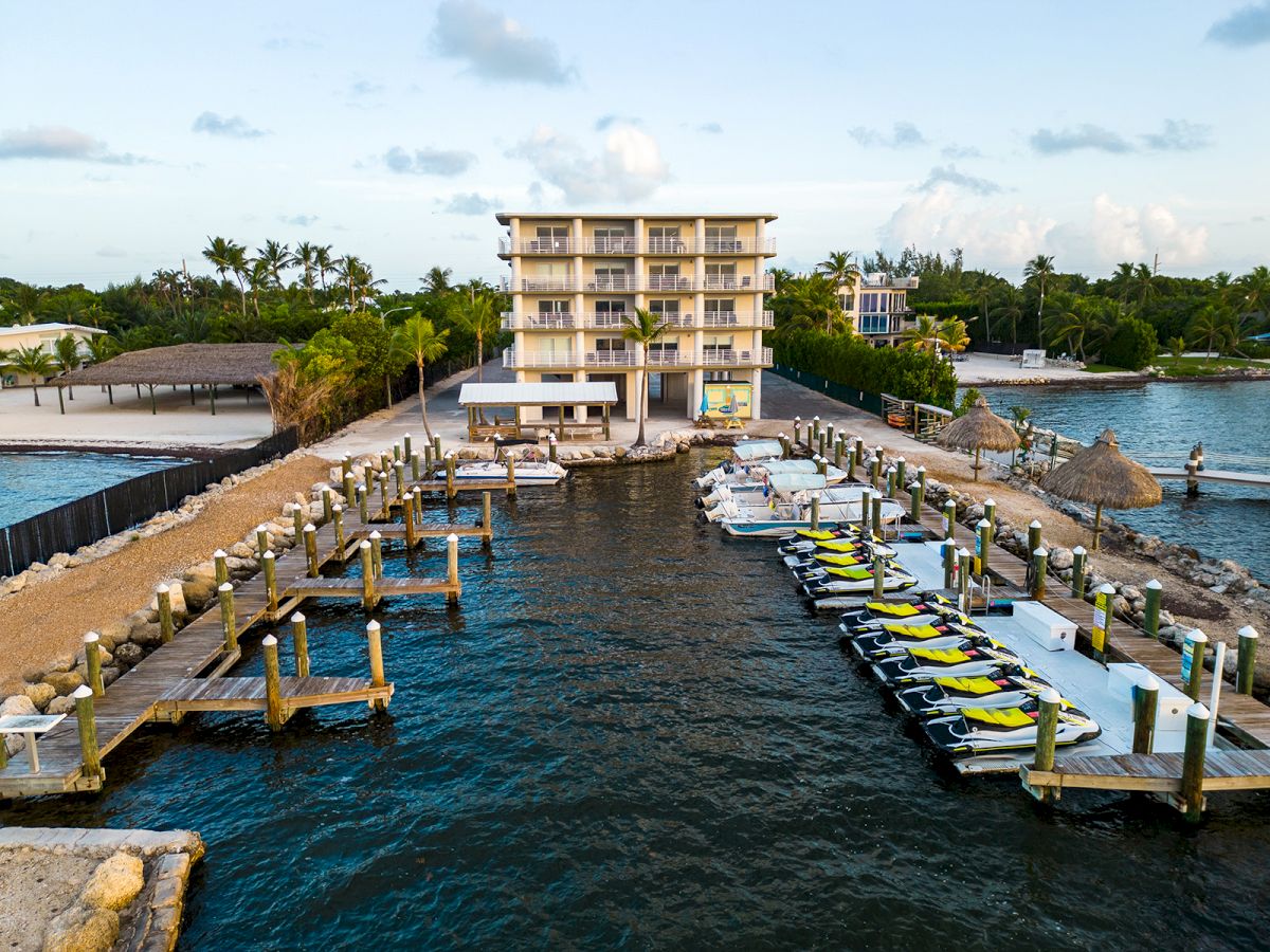 A beachfront resort with a multi-story building and docks packed with jet skis and boats on clear blue water.