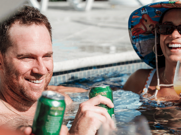 Two people are relaxing in a pool, holding drinks and smiling, with the woman wearing a sun hat and sunglasses.