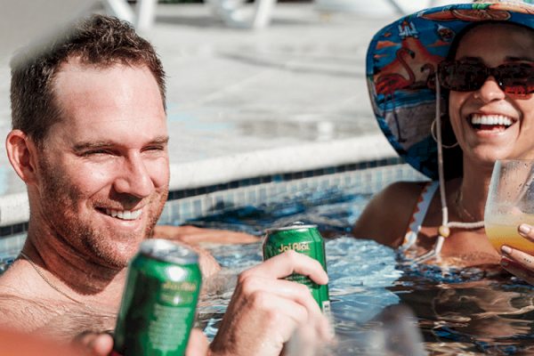 Two people are relaxing in a pool, holding drinks and smiling, with the woman wearing a sun hat and sunglasses.