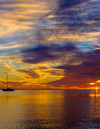 A sailboat is silhouetted against a vibrant sunset over calm water, with dramatic clouds and colorful reflections enhancing the scene.