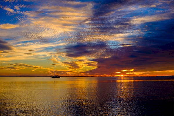 A sailboat on calm water is silhouetted against a vibrant sunset, with colorful clouds and reflections on the water's surface.