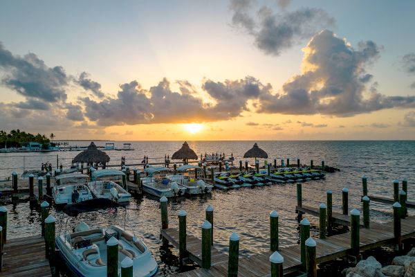 A beautiful sunset over a marina with boats docked at wooden piers and palm-thatched umbrellas in the background, clouds in the sky.