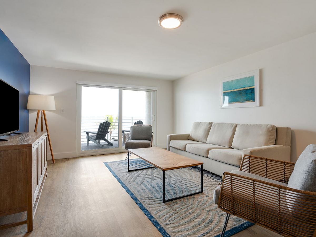 A modern living room with a beige sofa, wooden furniture, a TV, a blue accent wall, and sliding doors leading to a balcony with outdoor seating.