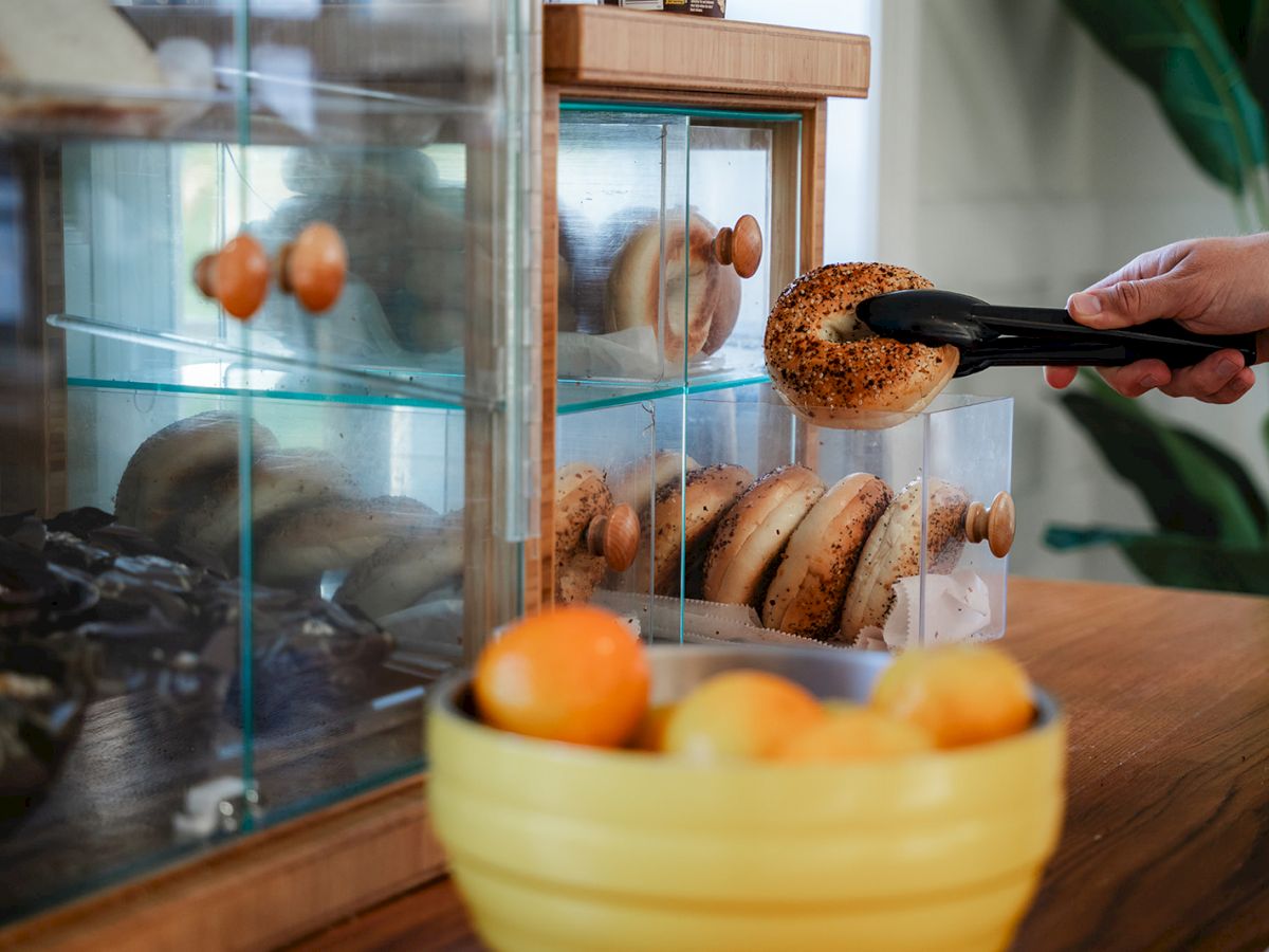 A person uses tongs to pick up a bagel from a glass display case on a wooden counter, with a bowl of oranges in the foreground.
