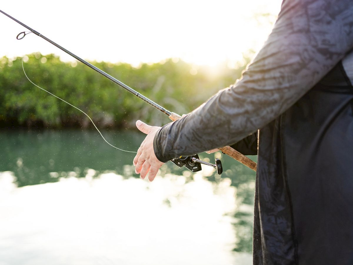 A person is holding a fishing rod near a body of water with greenery in the background, sunlight filtering through.