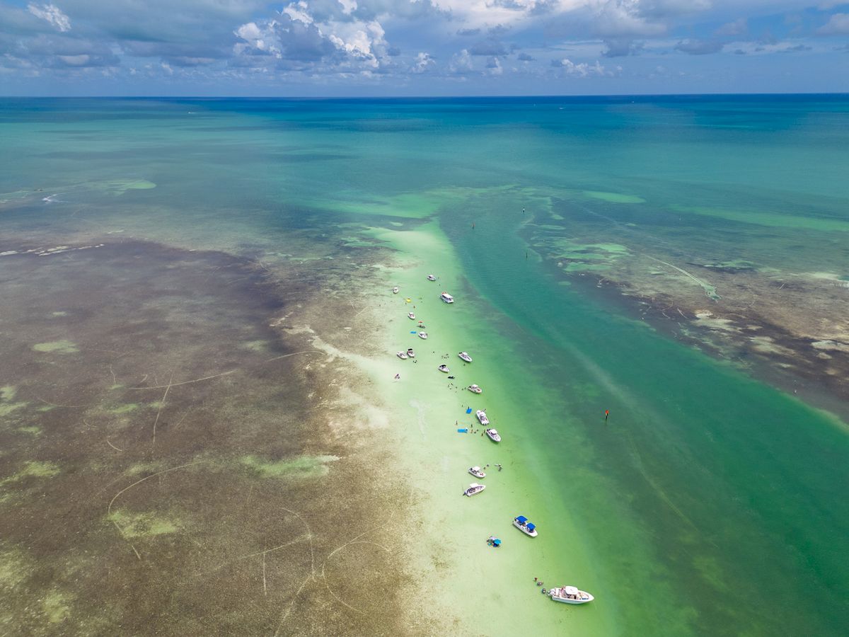 A serene aerial view of clear turquoise waters with several boats anchored along a sandy shoal amidst a tropical seascape under a partly cloudy sky.