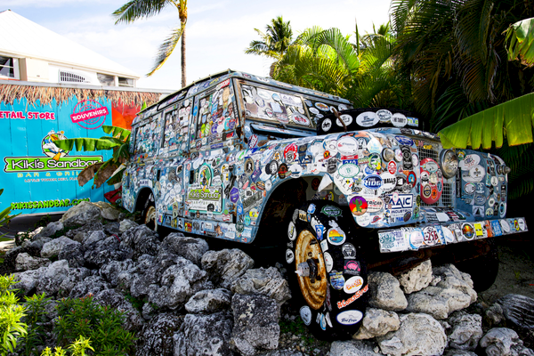 An old, sticker-covered vehicle is on rocks near lush greenery with a colorful sign that reads "Kiki's Sandbar."
