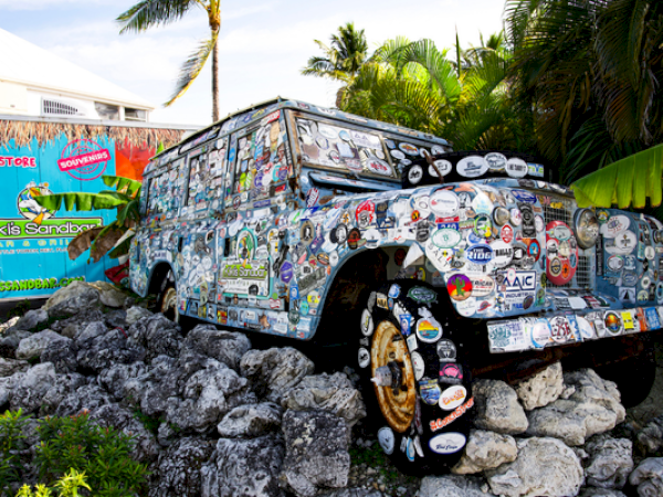 An old, sticker-covered vehicle is on rocks near lush greenery with a colorful sign that reads "Kiki's Sandbar."