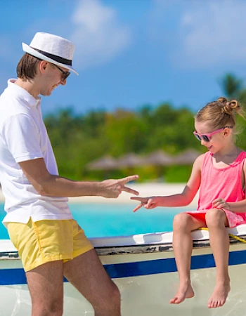 A man and a child are playing rock-paper-scissors on a boat by a tropical beach.