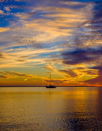 A sailboat on calm waters during a colorful sunset, with vibrant clouds and a reflection on the water's surface.