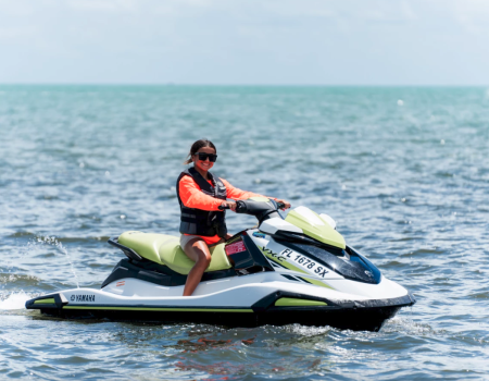 A person is riding a jet ski on the water, wearing sunglasses and a life jacket, under a clear sky.